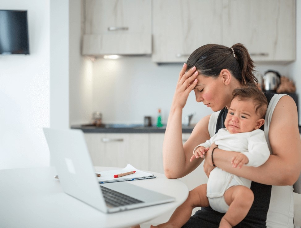 Madre cansada y estresada delante de un ordenador con un bebé en brazo que llora 